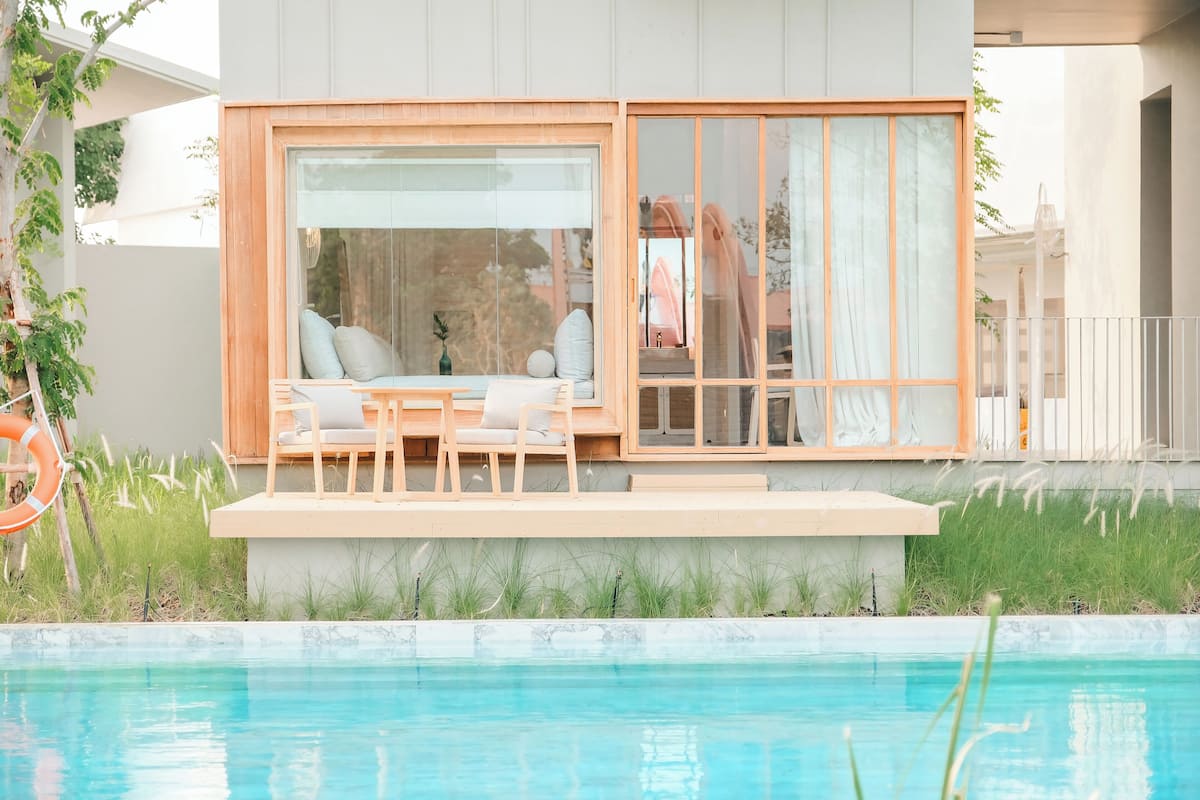 A modern outdoor seating area with a pool, featuring a wooden deck, cushioned chairs, and a glass enclosure.