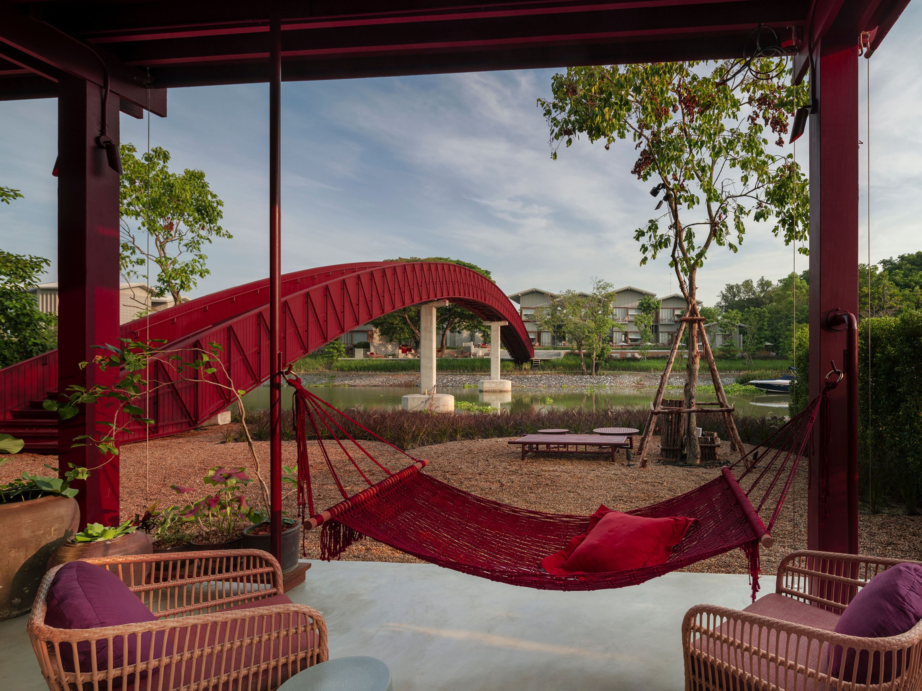 Un área de estar al aire libre techada con mobiliario de mimbre y una hamaca, con vistas a un cuerpo de agua y un puente rojo.