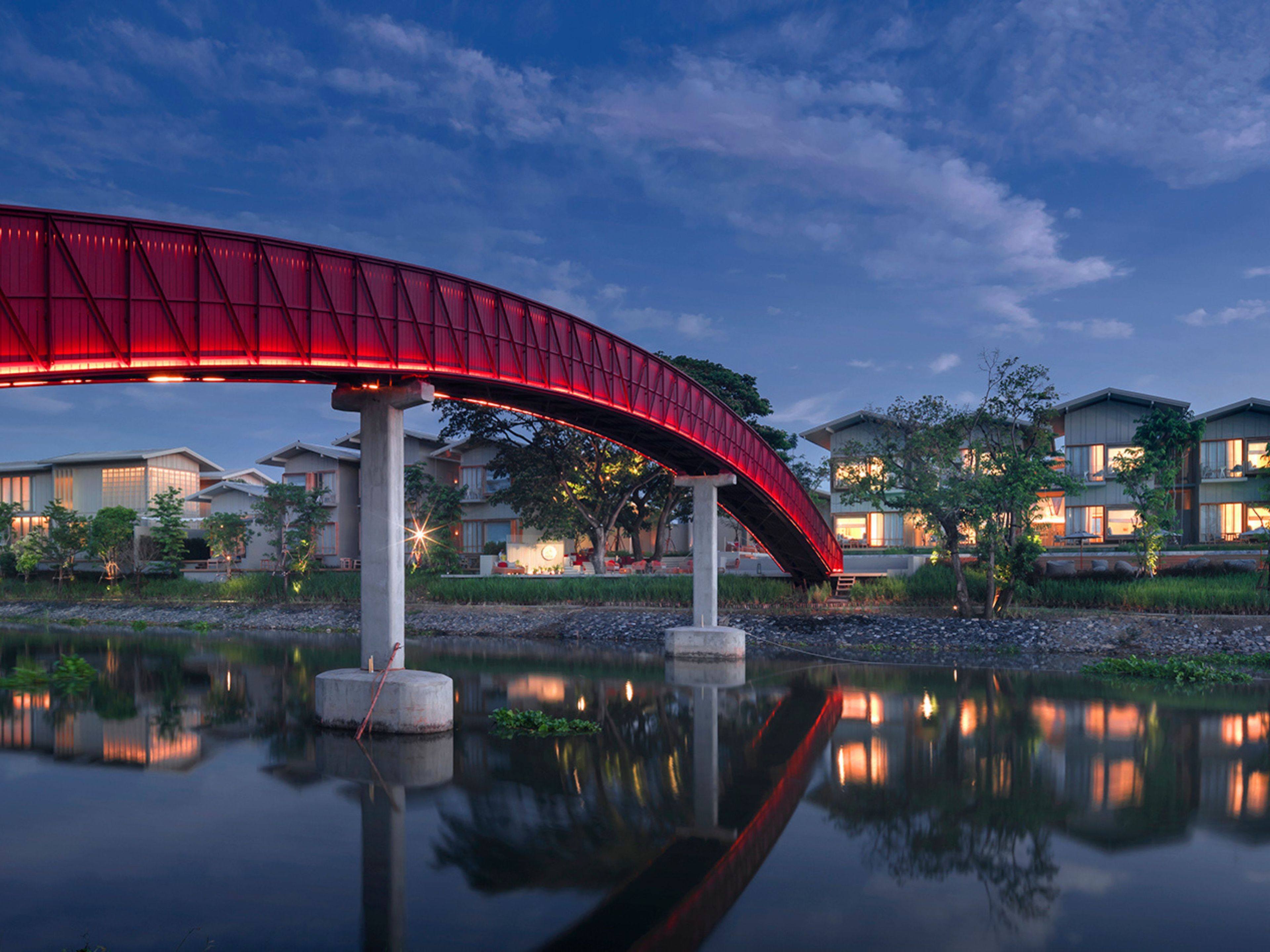 Uma ponte vermelha sobre águas calmas, com edifícios ao fundo.