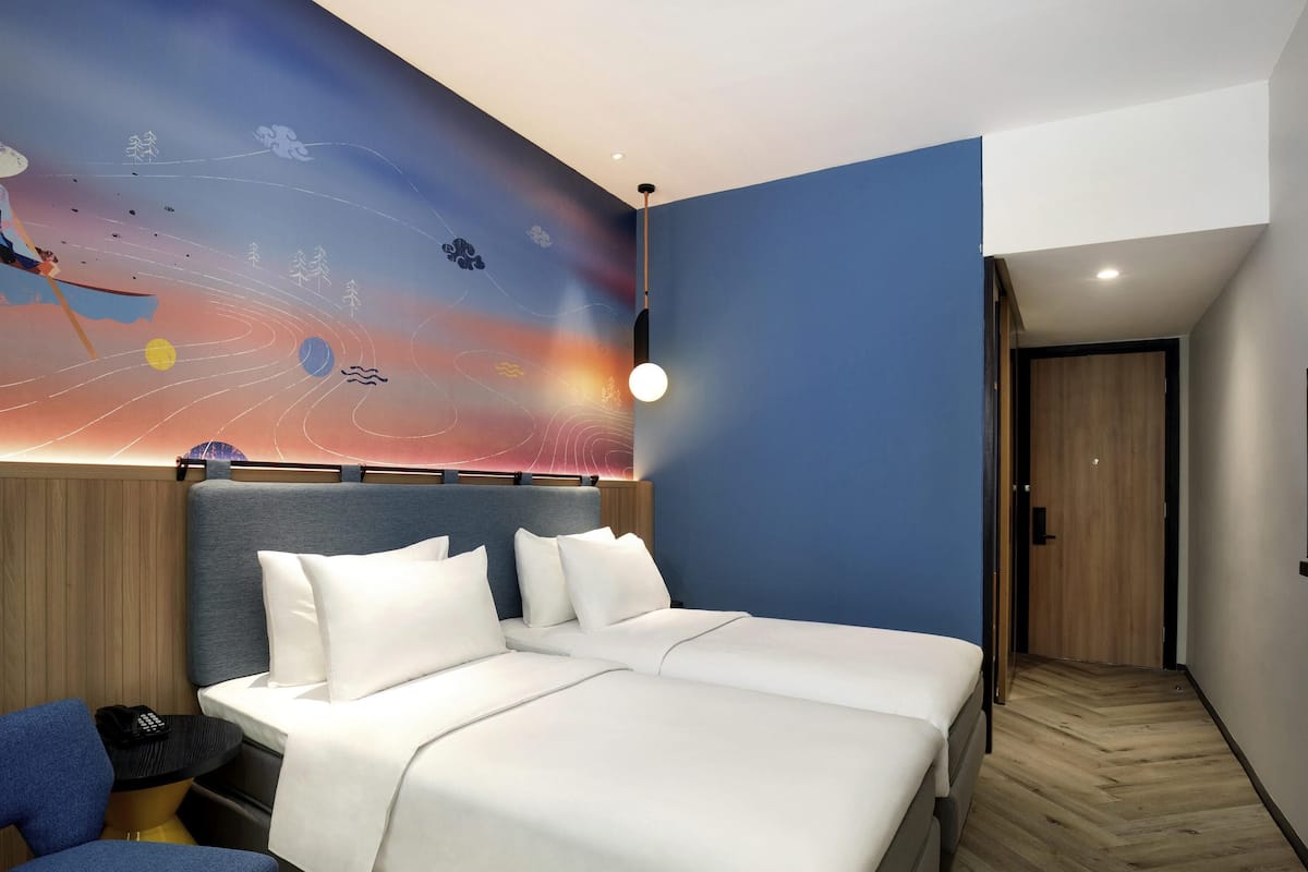 A modern hotel room with a large bed, a desk, and a chair. The room features a unique wall mural depicting a cosmic scene.