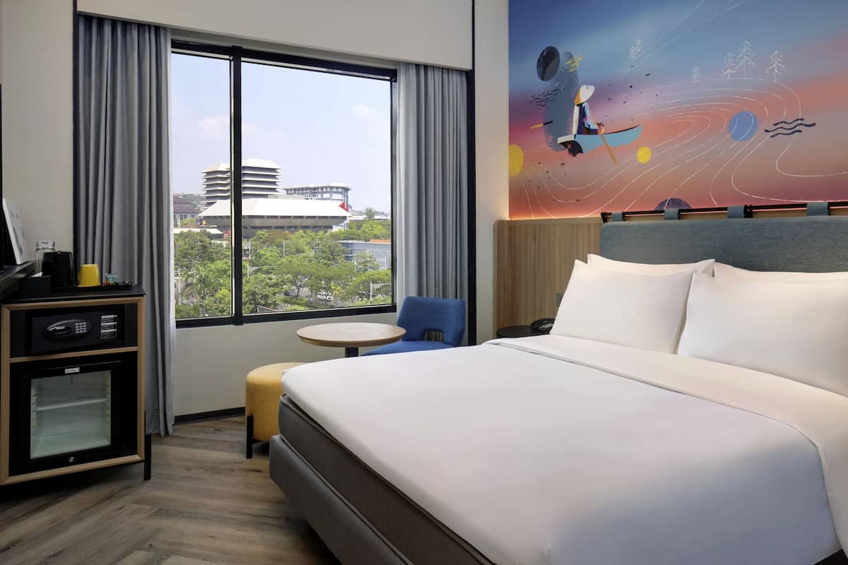 A hotel room with a large bed, a desk with a computer, a small table, and a view of the city through large windows.