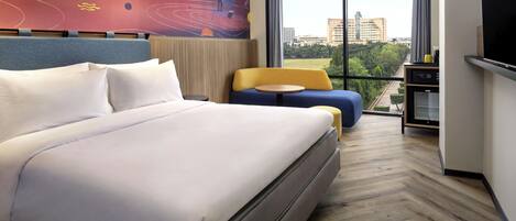 A modern hotel room with a large bed, a sofa, a small table, a TV, and a large window offering a city view.