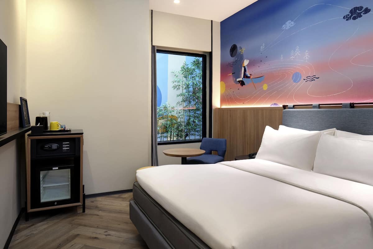 A modern hotel room with a large bed, a small desk with a coffee maker, a chair, and a wall mural depicting a space scene.