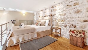 A bedroom with a stone wall, a bed with multiple pillows, a bedside table with a lamp, and a wooden bench. - Mouragia Loft - Waterfront Old Corfu Town (Corfu)