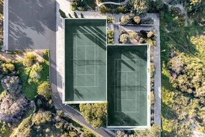 Luftaufnahme eines Tennisplatzkomplexes mit zwei Plätzen, umgeben von Grünflächen und einer Straße.