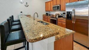 A kitchen with a granite countertop, wooden cabinets, stainless steel appliances, and a black leather bar stool.