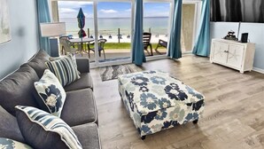 A room with a sofa, ottoman, and a view of the beach.