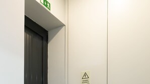 Un pasillo con una puerta cerrada, una señal de salida verde y un letrero de advertencia sobre riesgos eléctricos.