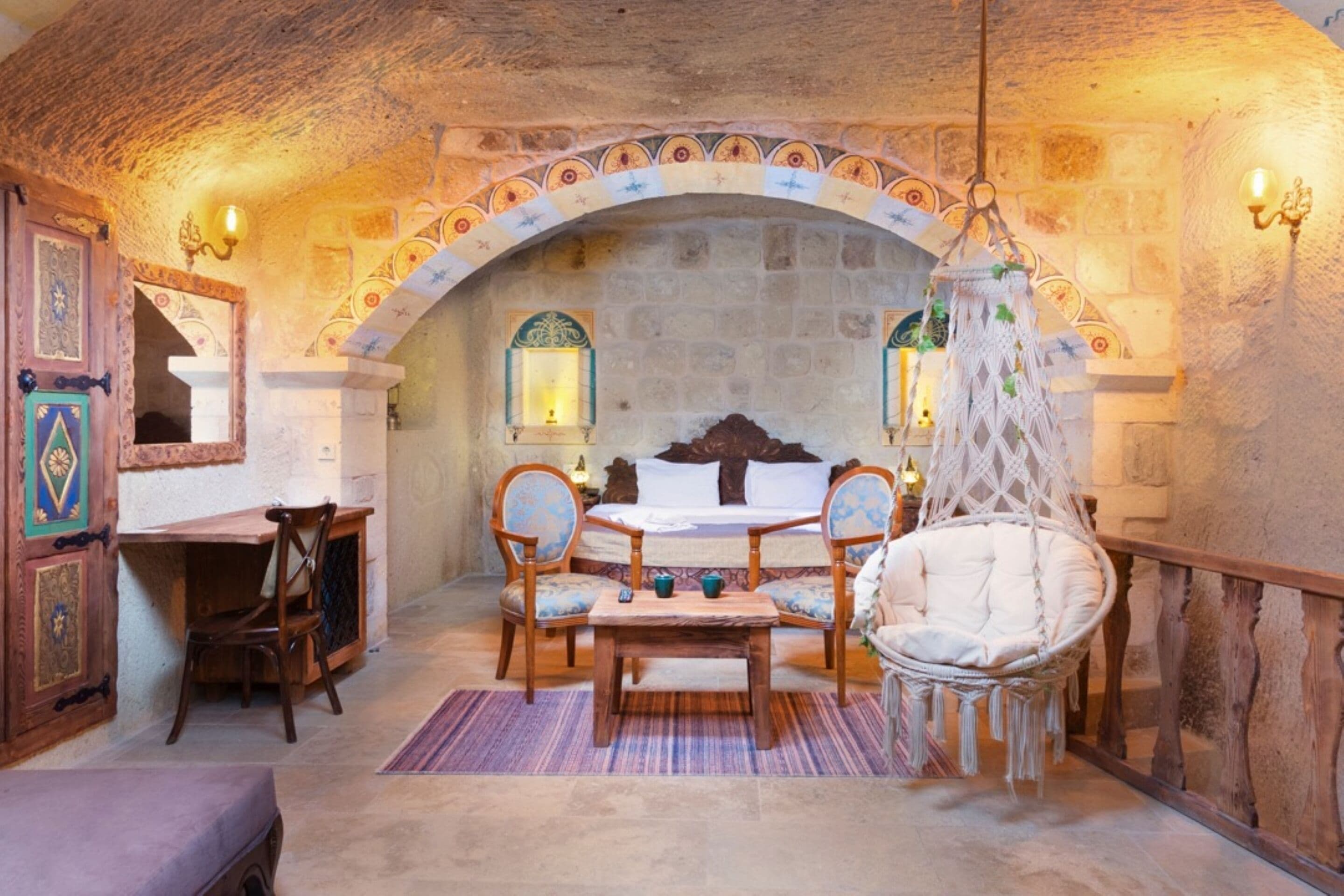A cozy room with a stone archway, a bed with a wooden headboard, a hanging chair, and a small table with chairs.