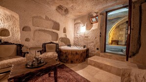 A stone-walled room with a wooden bathtub, two upholstered chairs, a coffee table, and a view of an adjacent room through a glass door. - Capp Royal Cave Hotel (Ürgüp)