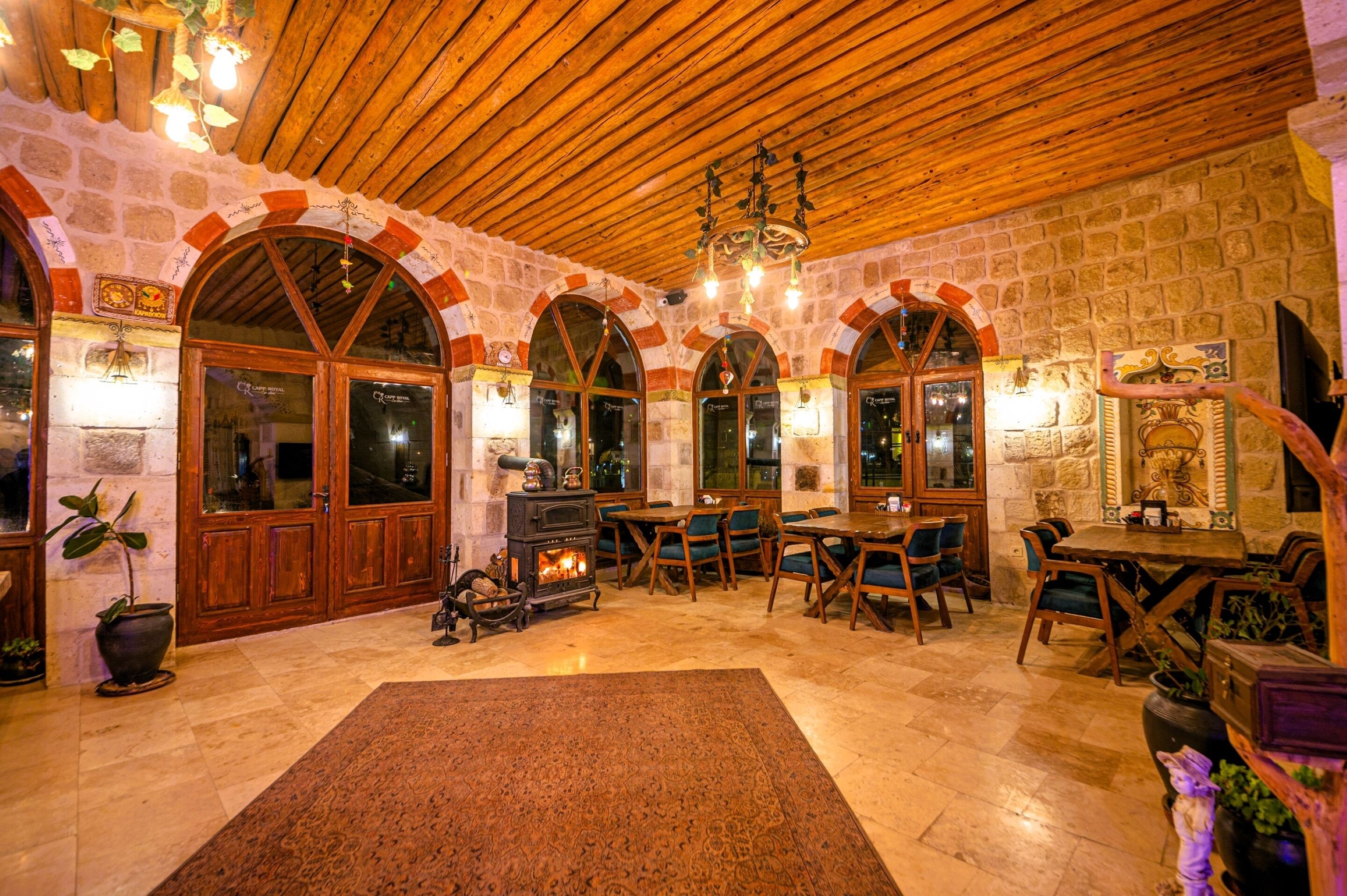 A cozy interior with stone walls, wooden beams, and arched windows. There is a fireplace, wooden furniture, and a rug on the floor.