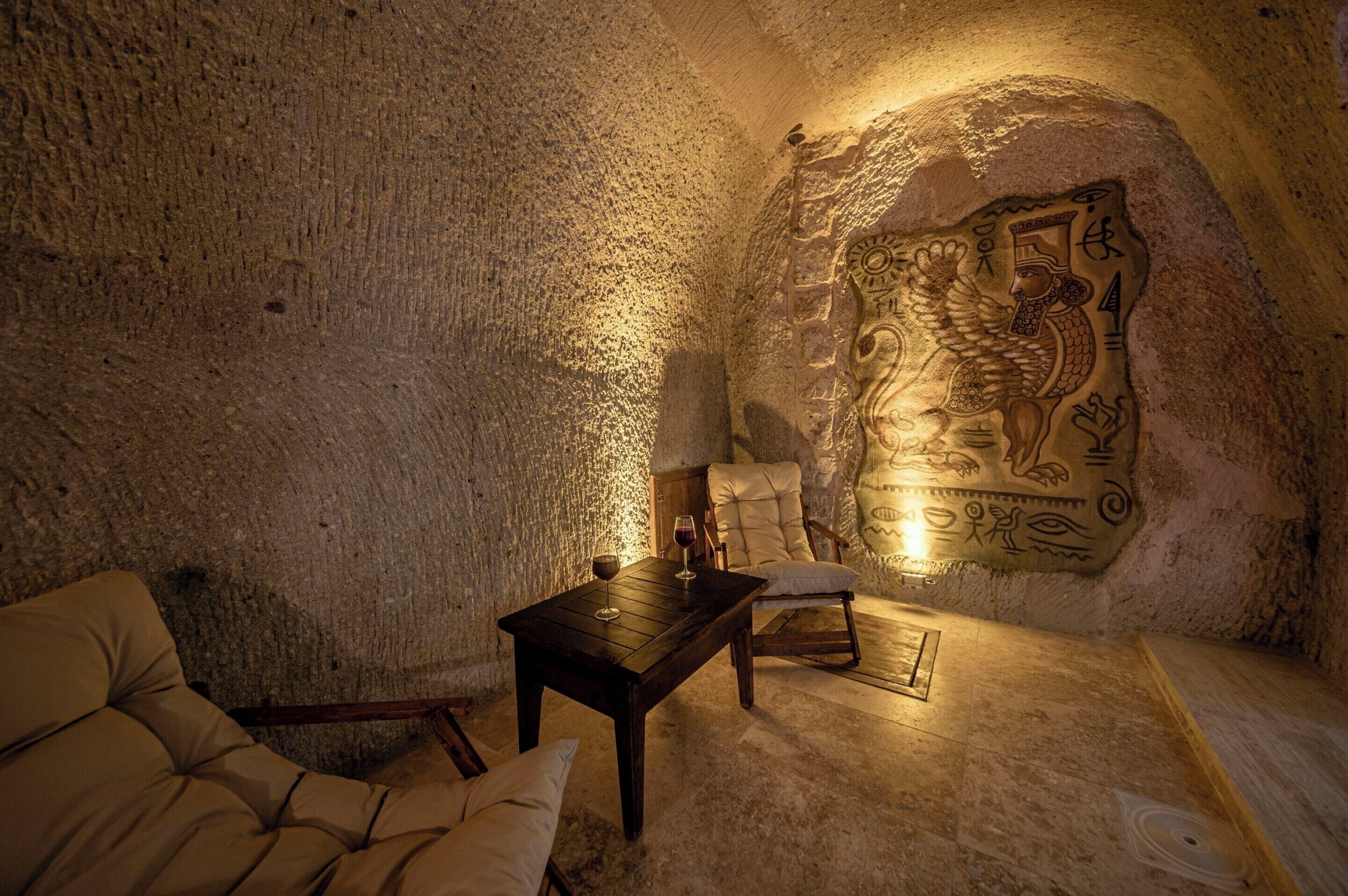 A cozy interior with a stone wall, a carved mural, a wooden table with a glass, and a cushioned chair.