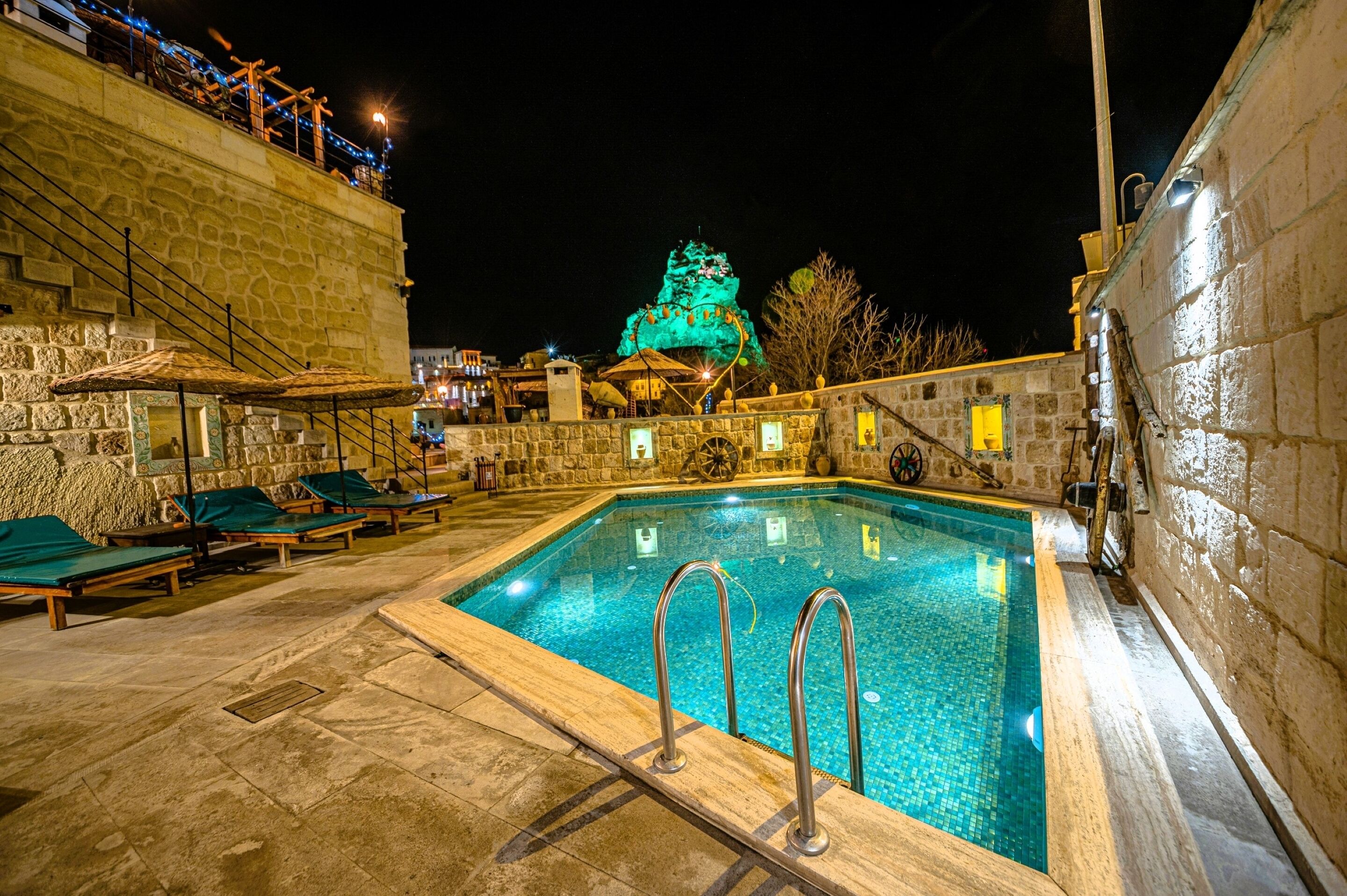 Una piscina con sillas de descanso, un muro de piedra y un edificio con una estructura iluminada en verde al fondo.