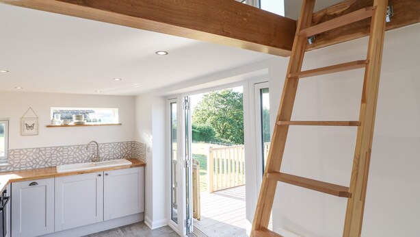 Une cuisine moderne avec des armoires blanches, un plan de travail en bois et un évier encastré. On y trouve une échelle en bois appuyée contre le mur et une porte-fenêtre coulissante donnant sur une terrasse extérieure.