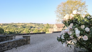 Un sentier de pierres mène à un bâtiment en pierre, d’où l’on peut admirer des collines verdoyantes et un arbre aux fleurs roses.