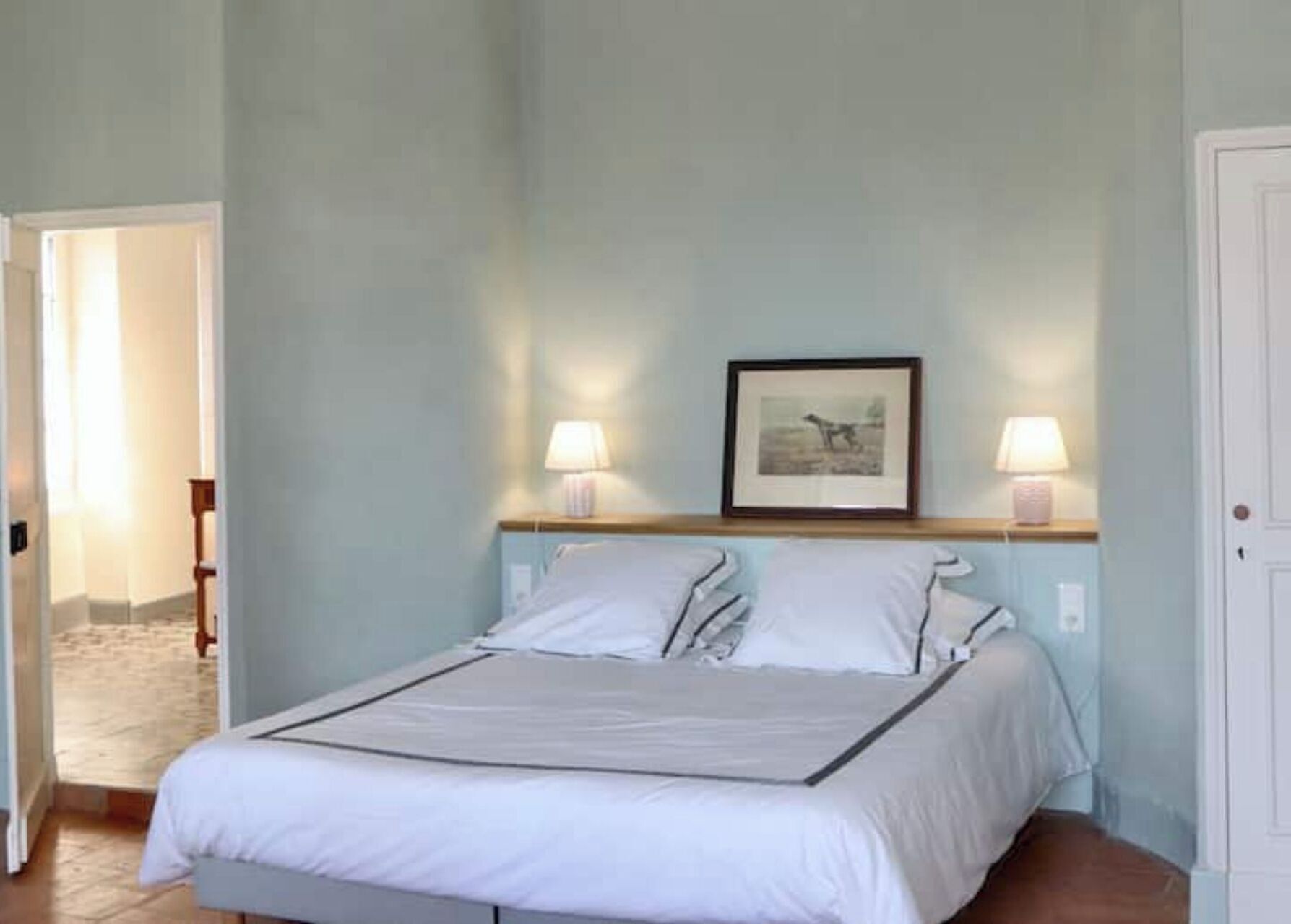 A bedroom with a bed, two bedside lamps, a framed picture on the wall, and a door to the right.