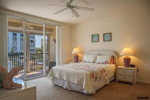 Ein Schlafzimmer mit Bett, zwei Nachttischlampen, einem Deckenventilator und einem Balkon mit Blick auf Gebäude.