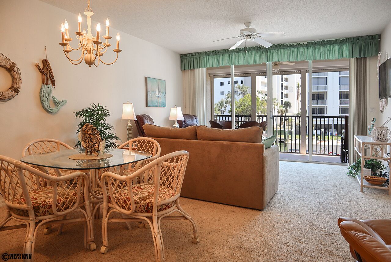 A living room with a glass-top dining table, wicker chairs, a brown sofa, and a balcony with a view of buildings.