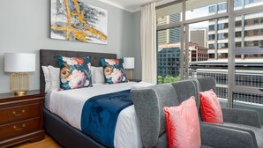 A modern hotel room with a large bed, grey sofa, and a city view through large windows.