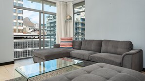 A modern living room with a grey sofa, a glass coffee table, and a large window offering a city view.