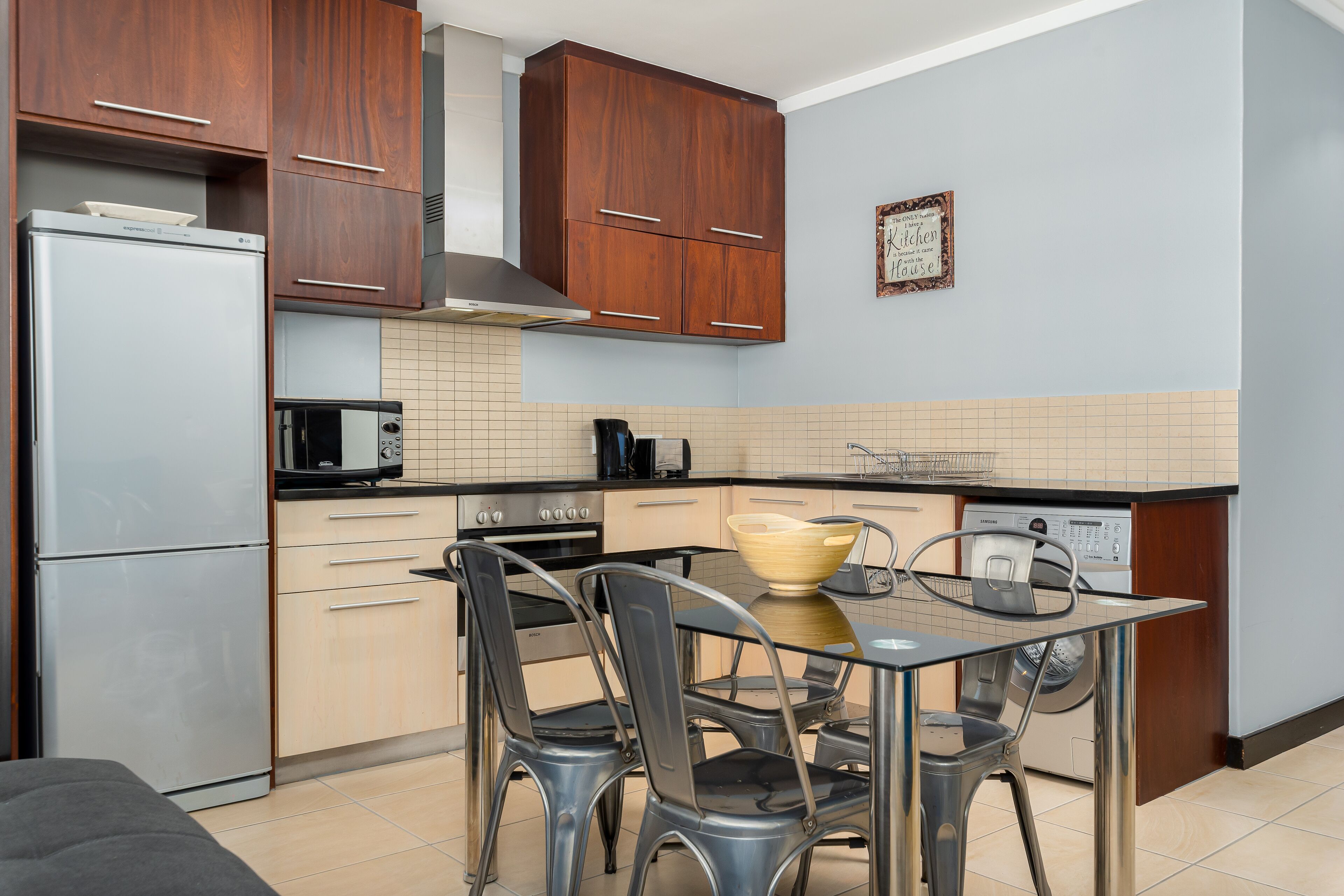 A compact kitchen with wooden cabinets, a stainless steel refrigerator, a microwave, a stove, and a dining table with chairs.