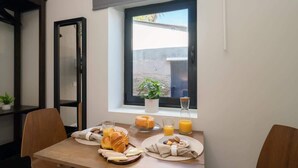 A breakfast table with croissants, a fruit plate, and juice, near a window with a view of a building and a plant. - Cozy Tiny House | Prime Location by HostWise (Porto)