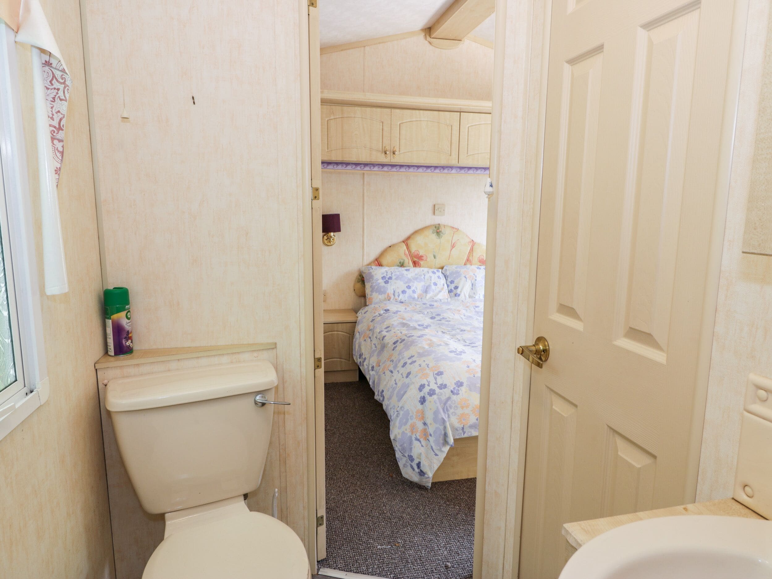 A narrow hallway leading to a bedroom with a bed, a toilet, and a window with curtains.