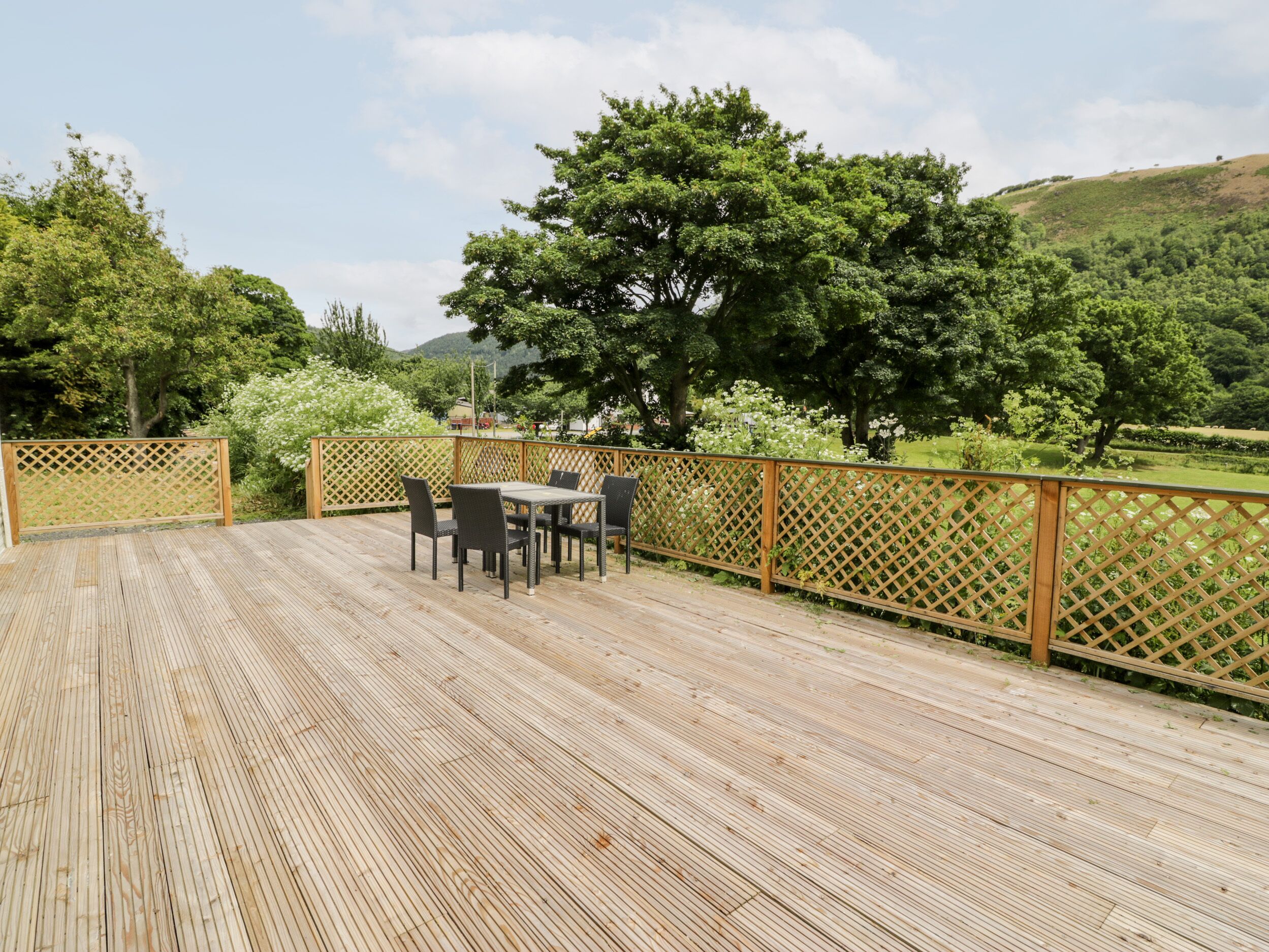 Eine Holzterrasse mit Tisch und Stühlen, umgeben von einem Holzzapfen, mit Blick auf eine grüne Landschaft mit Bäumen und Hügeln.