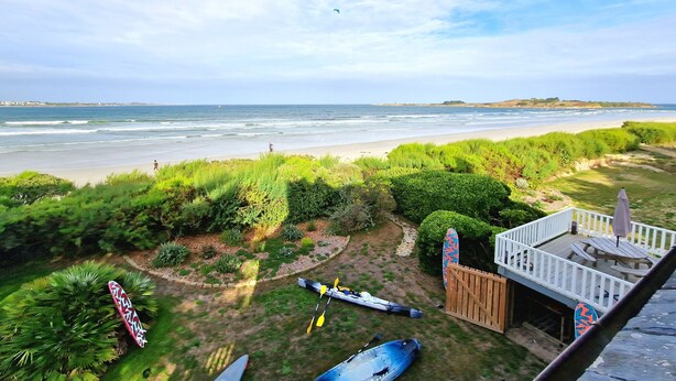 A view of a beach with kayaks and a paddleboard, a wooden deck with a table and chairs, and a grassy area with bushes. - Dream destination, right by the sea, (Santec)