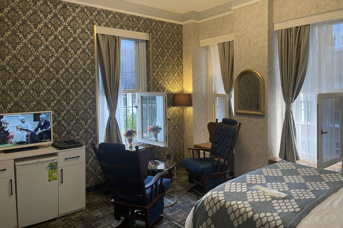 A hotel room with a bed, a desk with a television, a chair, a lamp, and a window with curtains.