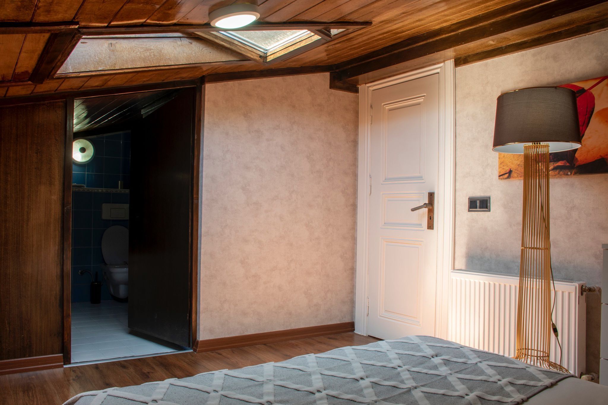 A bedroom with a wooden ceiling, a bed with a patterned bedspread, a white door, and a floor lamp.
