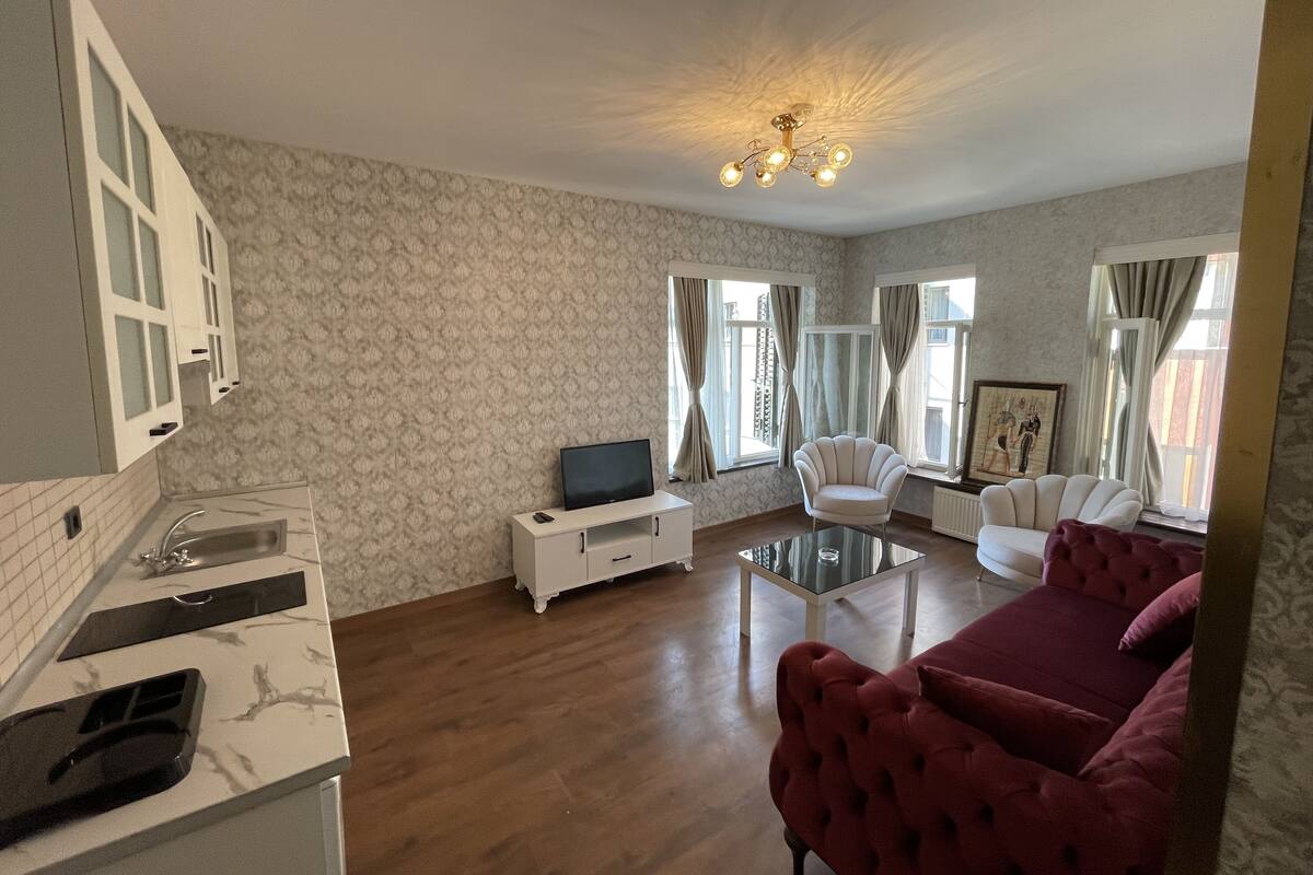 A living room with a TV, white armchairs, a red sofa, a glass coffee table, and a patterned wallpaper.