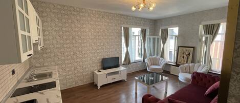 A living room with a TV, white armchairs, a red sofa, a glass coffee table, and a patterned wallpaper.