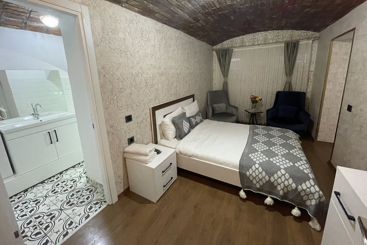 A bedroom with a bed, a nightstand, a chair, a small table with a vase, and a bathroom with a sink and mirror.
