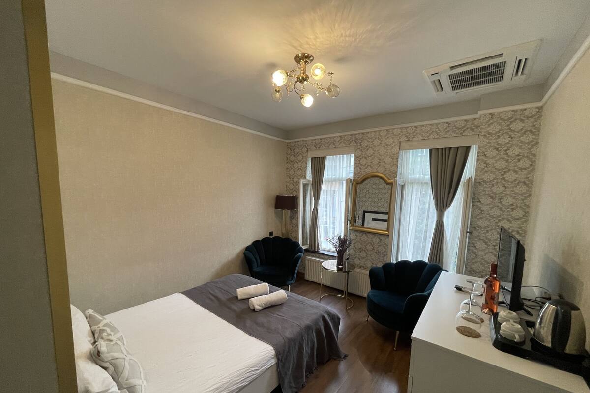 A hotel room with a bed, two chairs, a desk, a mirror, and a chandelier.