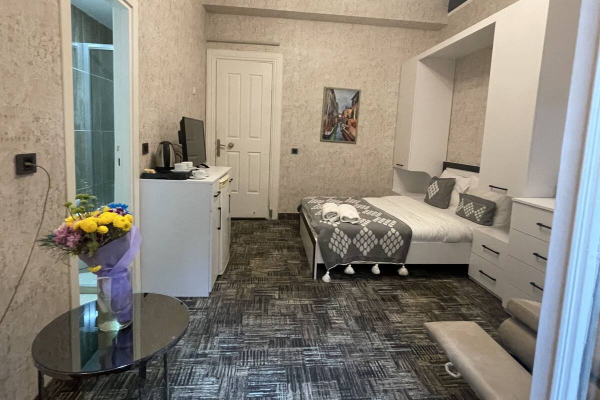 A compact hotel room with a bed, a desk with a computer, a small table with a vase of flowers, and a mirror.