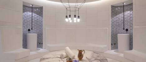 A modern spa room with a circular marble table, two white towels, a copper pitcher, and a small bowl of bath salts.