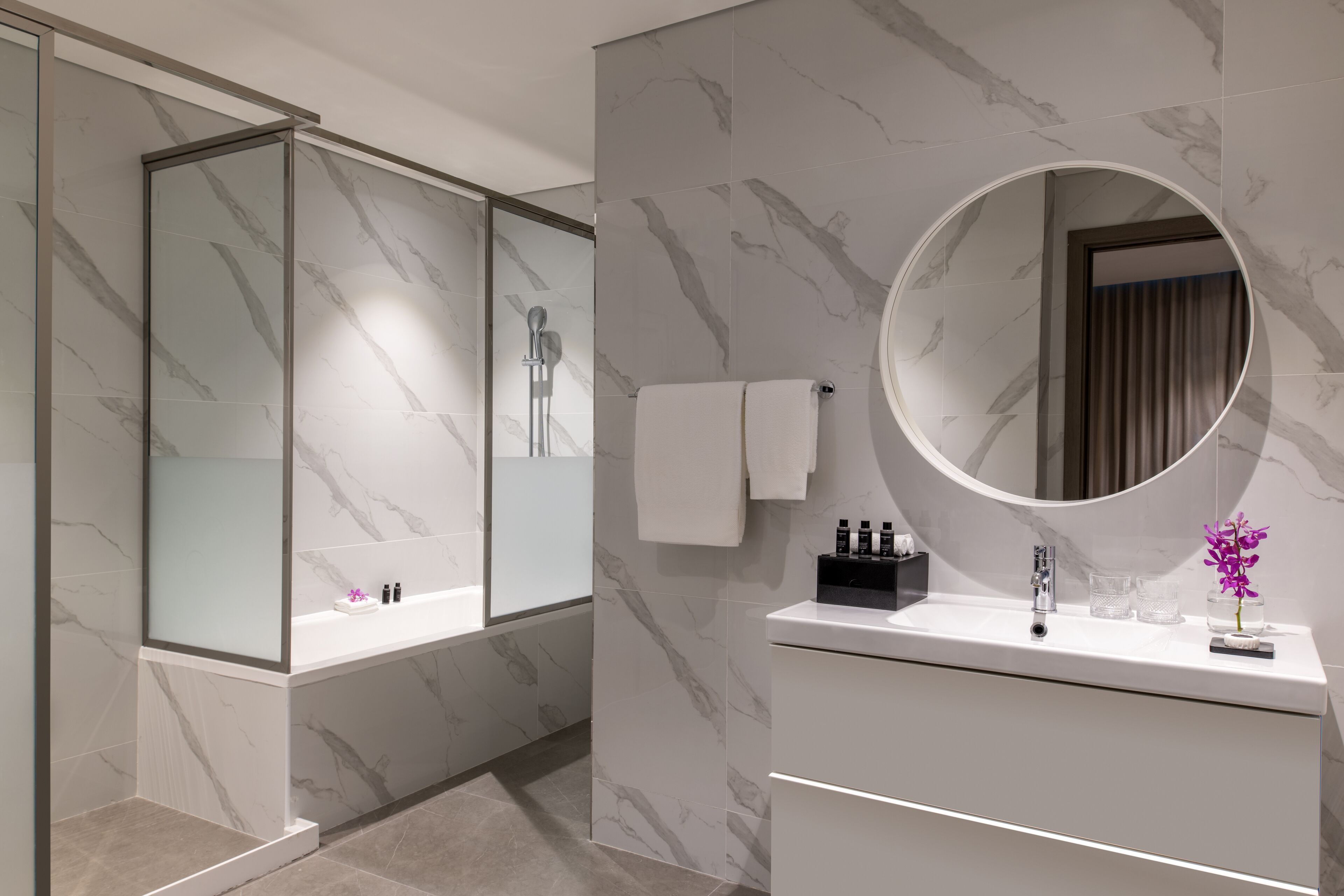 A modern bathroom with a glass-enclosed shower, a round mirror, and a white vanity with toiletries.