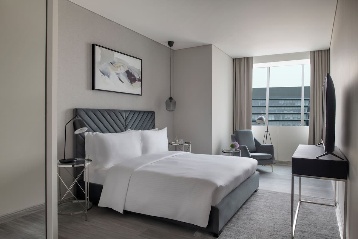 A modern hotel room with a large bed, a desk, a chair, and a view of the city.