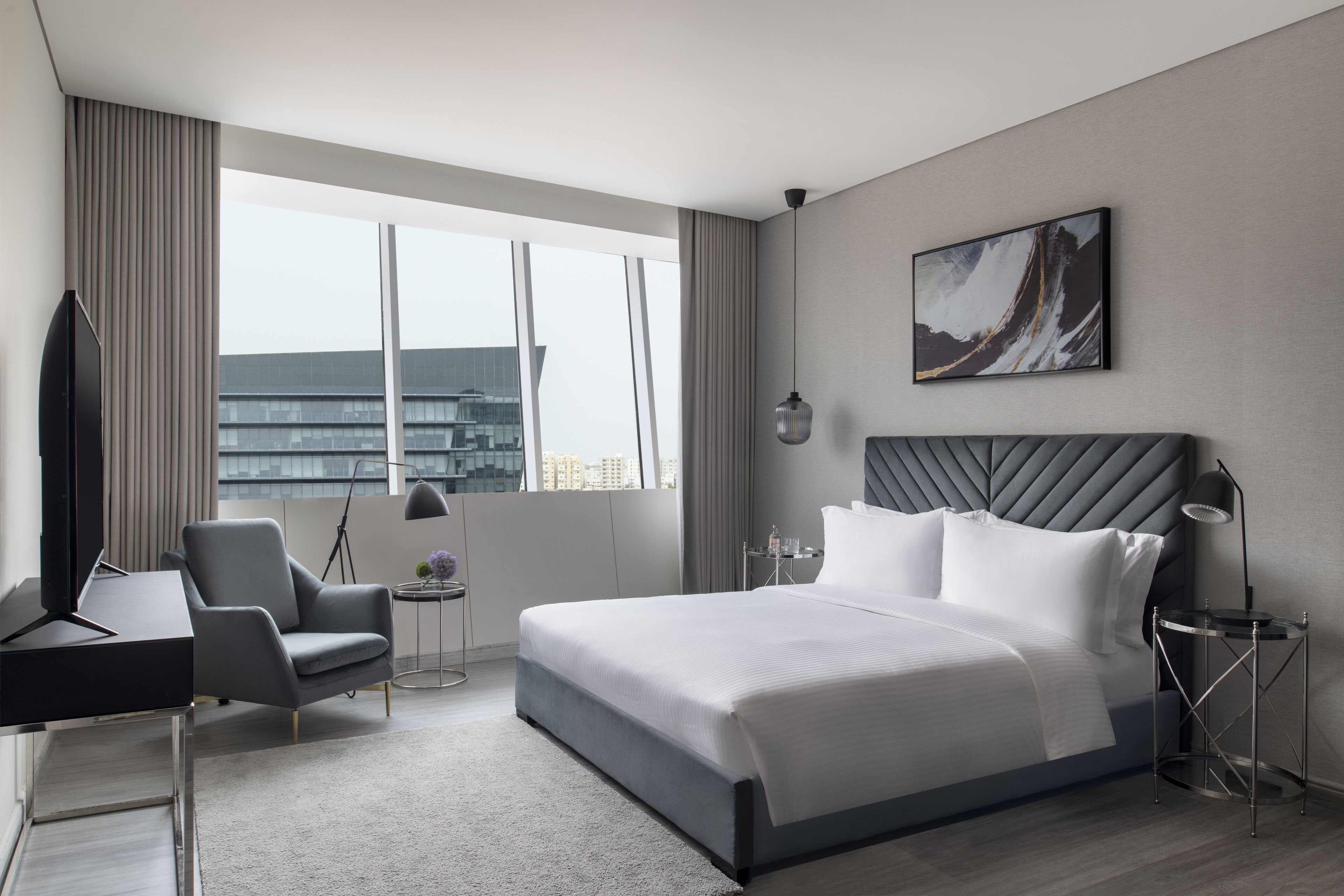 A modern hotel room with a large bed, a desk, a chair, a side table, a lamp, and a painting on the wall.