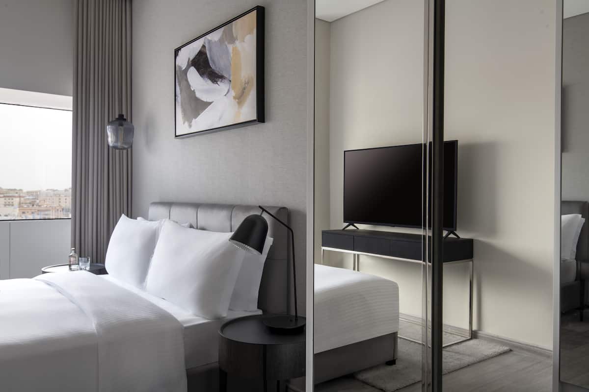 A modern hotel room with a large bed, a flat-screen TV, a bedside table with a lamp, and a city view through the window.