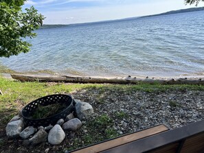 A lakeside view with a wooden dock, a black basket with rocks, and a grassy area. - Direct Lake Leelanau sunset view, fire pit, grill, dock, boat lift, dog ok (Traverse City)