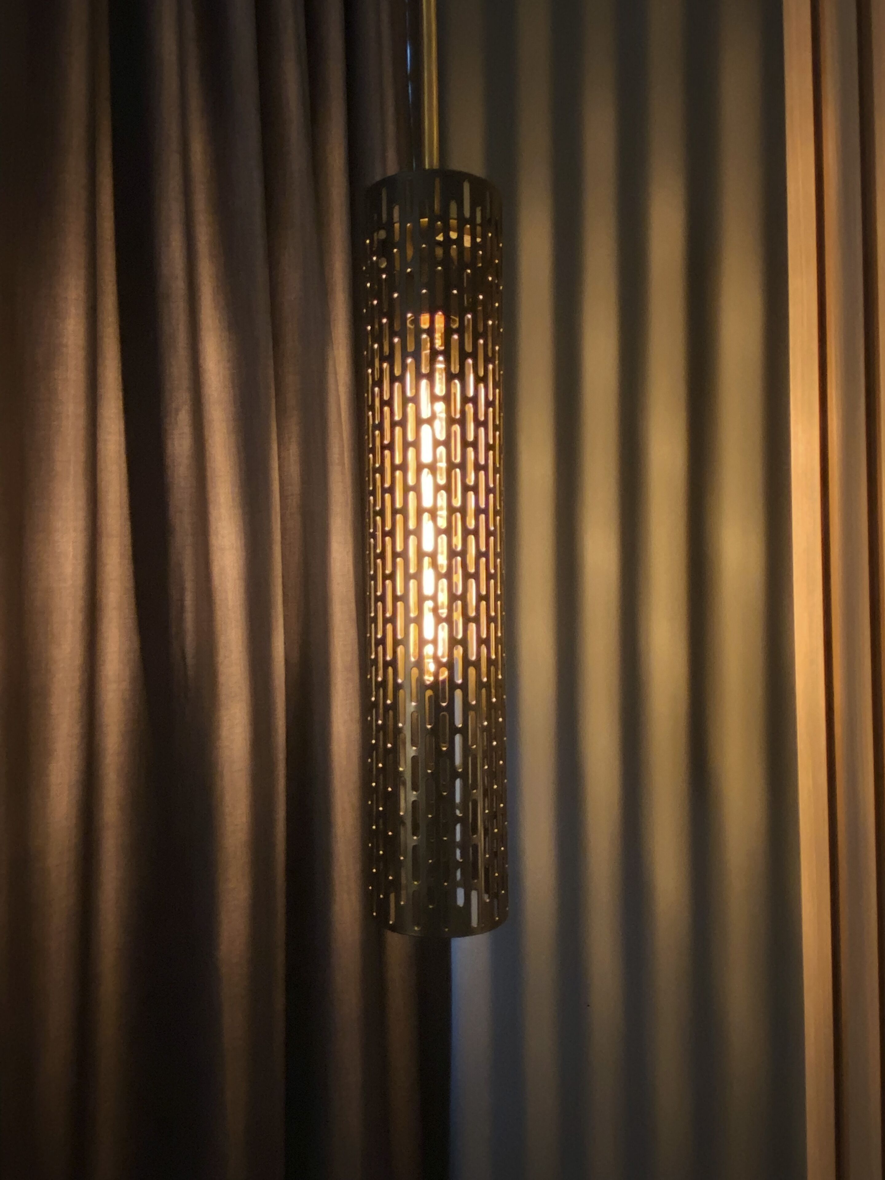 Una lampada cilindrica con decorazioni in metallo forato, sospesa di fronte a tende scure.