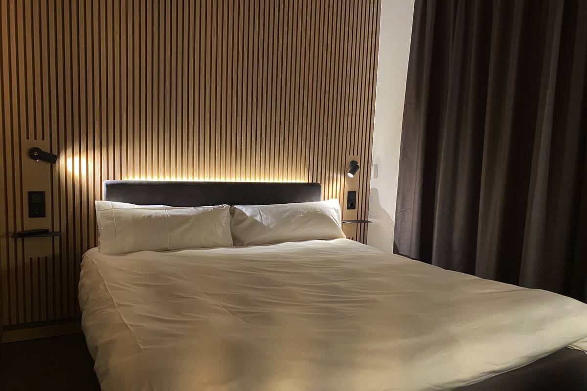 A hotel room with a bed, a headboard, a nightstand, and a wall with vertical lines.