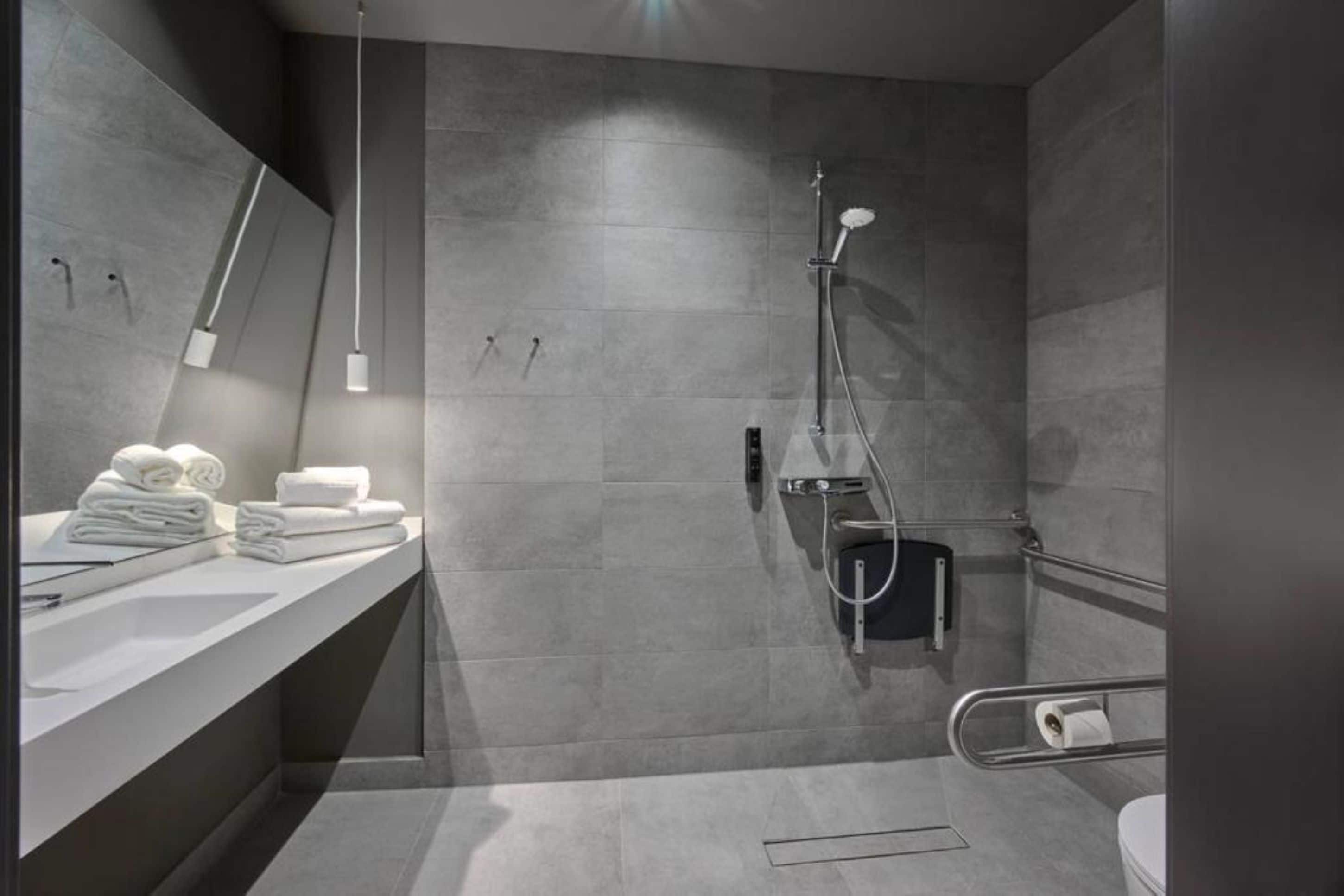 Een moderne badkamer met een douchebekleding van grijze tegels, een witte wastafel met handdoeken en een handgreep.