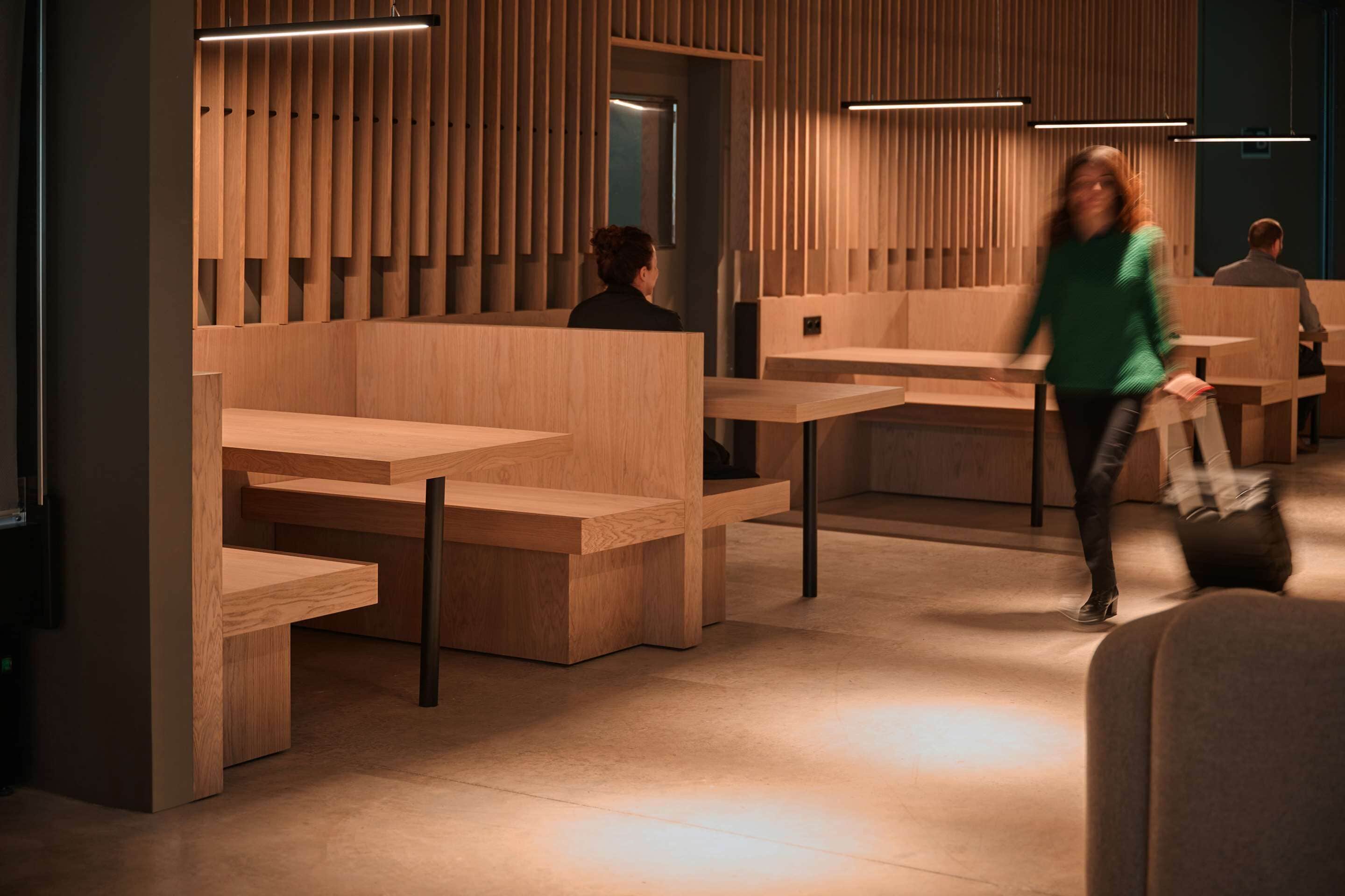 Un interior moderno con separadores y mesas de madera, una persona caminando y otra sentada al fondo.