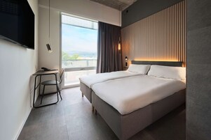 Ein modernes Hotelzimmer mit einem großen Bett, einem Schreibtisch mit Stuhl, einem an der Wand befestigten Fernseher und Blick ins Freie.