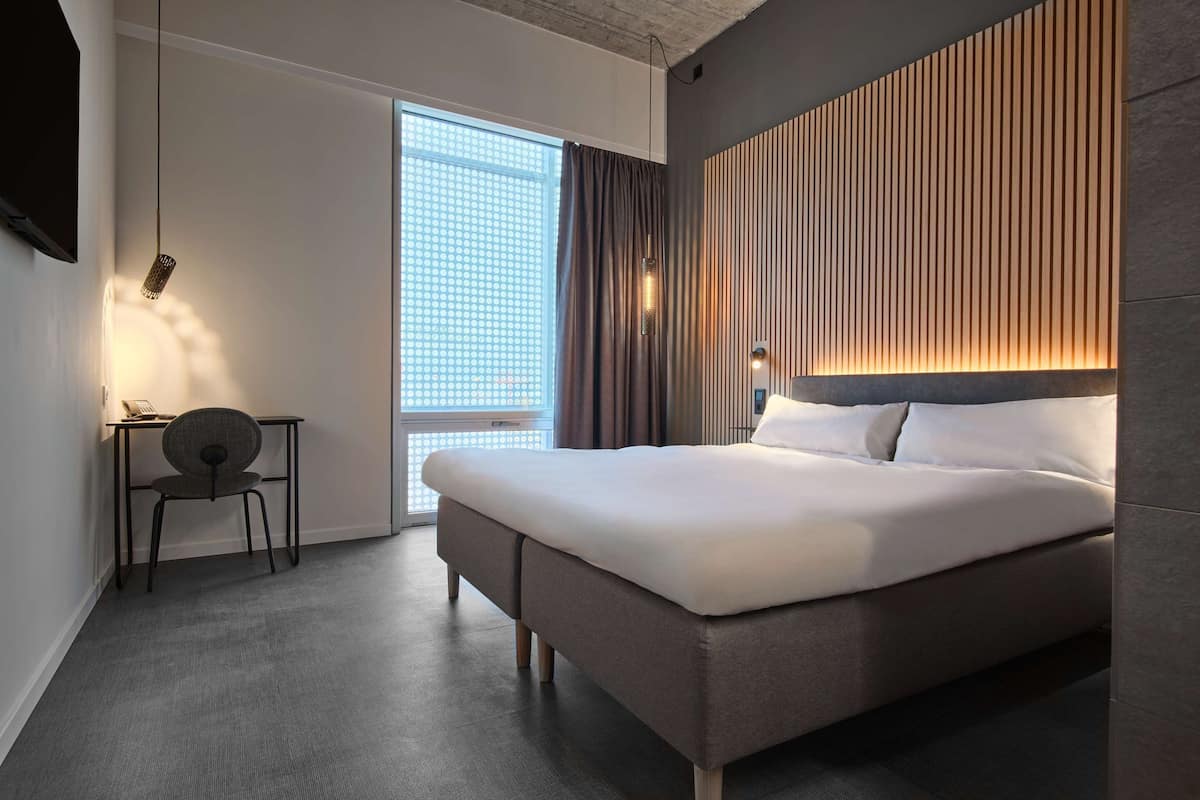 A modern hotel room with a large bed, a desk, and a chair. There is a window with curtains, a wall-mounted light, and a TV on the wall.