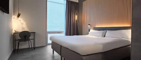 Une chambre d’hôtel moderne avec un grand lit, un bureau et une chaise. On y trouve une fenêtre avec des rideaux, une lampe fixée au mur et un téléviseur mural.
