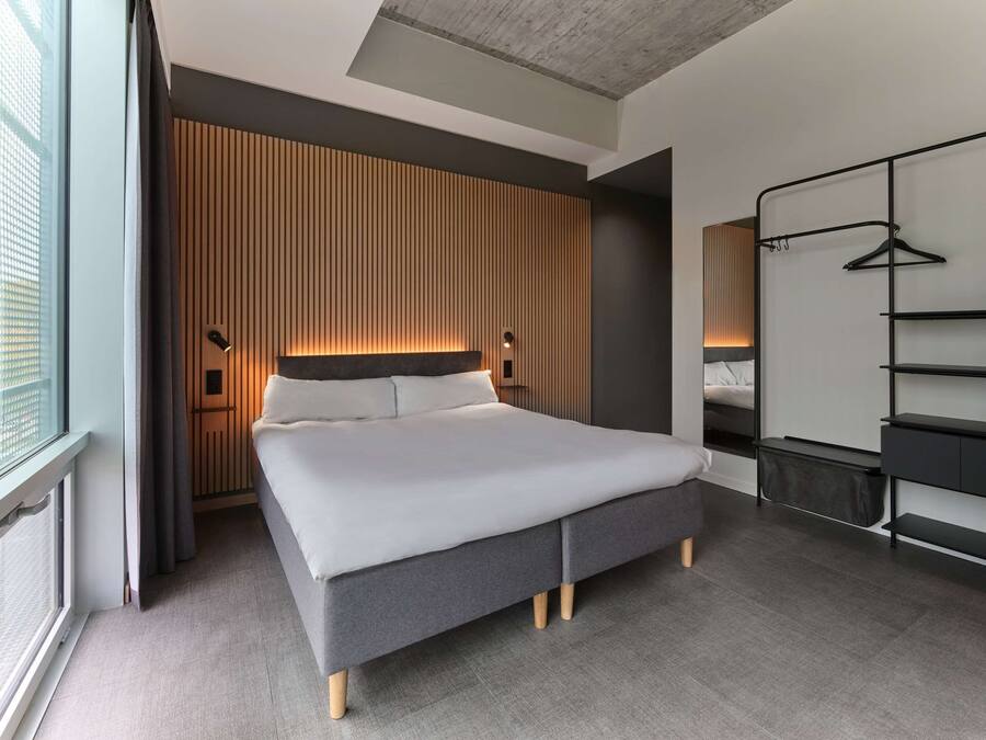 A modern hotel room with a large bed, a wardrobe with shelves, a bedside table, and a window with curtains.